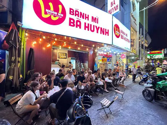 Diners sitting at small sidewalk tables outside the brightly illuminated Bánh Mì Bà Huynh eatery at night.
