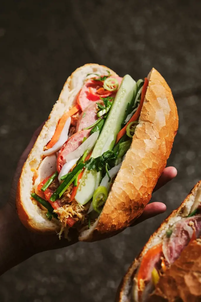 A hand holding a fully stuffed Vietnamese bánh mì sandwich filled with meats, fresh cucumber, cilantro, and chili peppers in a crusty baguette.
