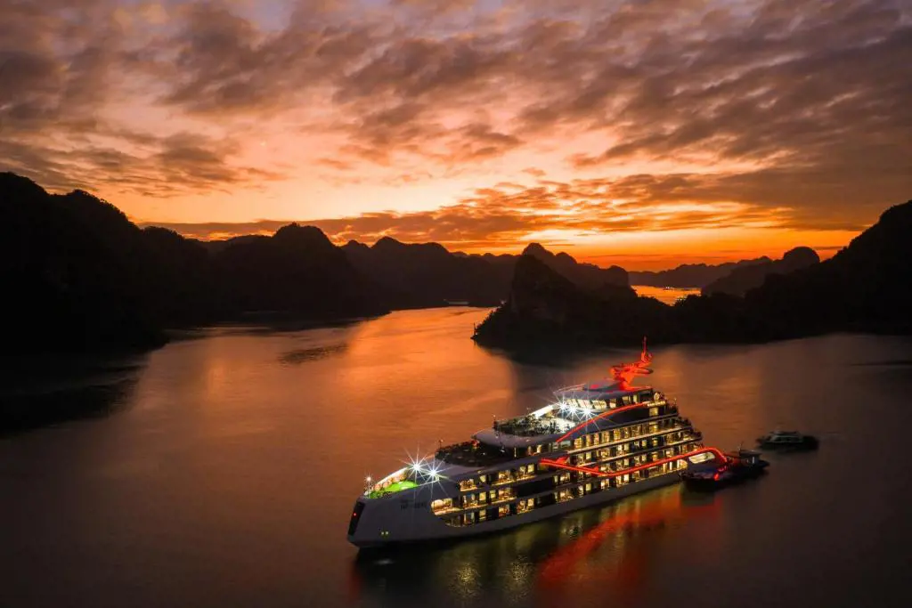 Aerial view of a brightly lit multi-deck luxury cruise ship anchored on calm bay waters surrounded by limestone karst mountains at sunset.