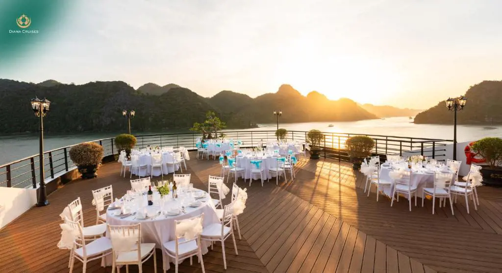 White round banquet tables and chairs arranged on a wooden cruise ship deck overlooking limestone mountains at sunset.