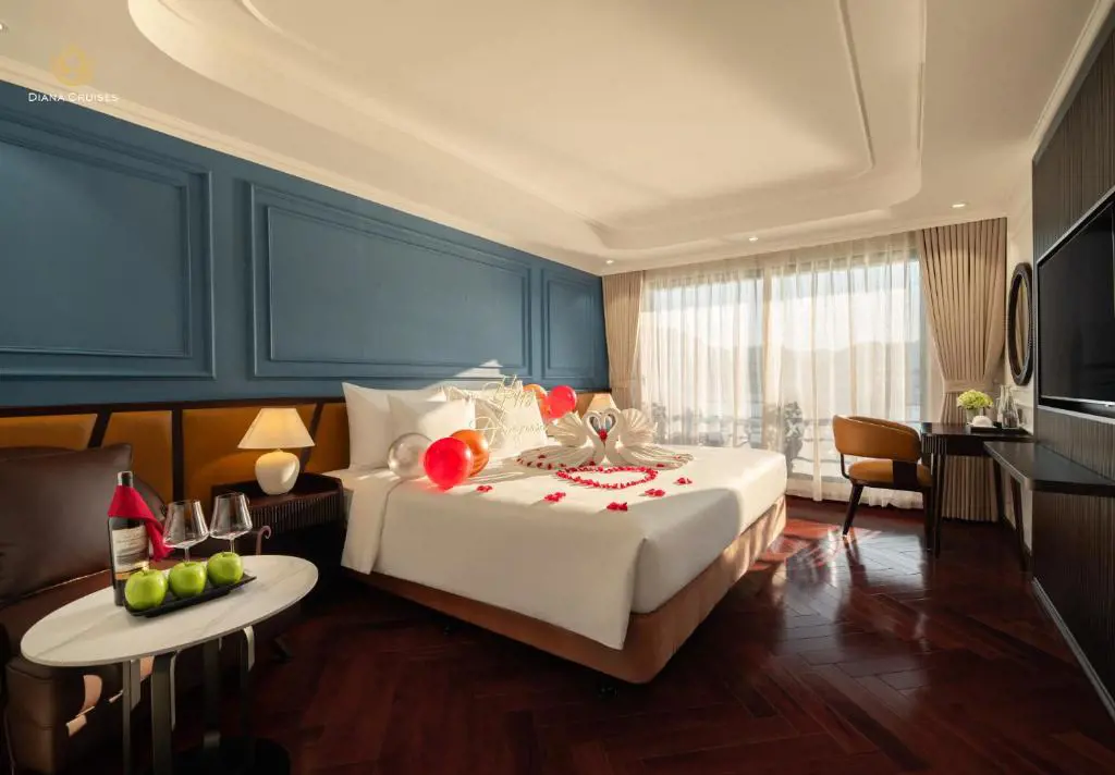 Luxury cruise cabin bedroom with a king bed decorated with swan-folded towels, red rose petals, and balloons beside a scenic balcony view.