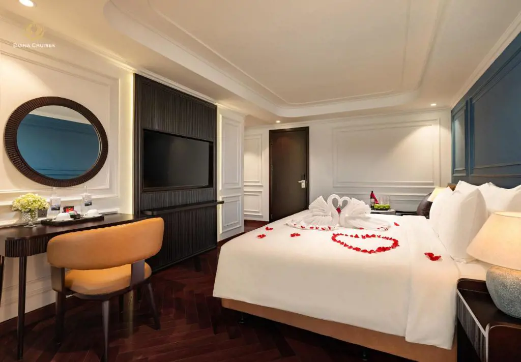 A luxury cruise cabin bedroom featuring a bed decorated with romantic towel swans and rose petals, a vanity mirror, and dark wood flooring.