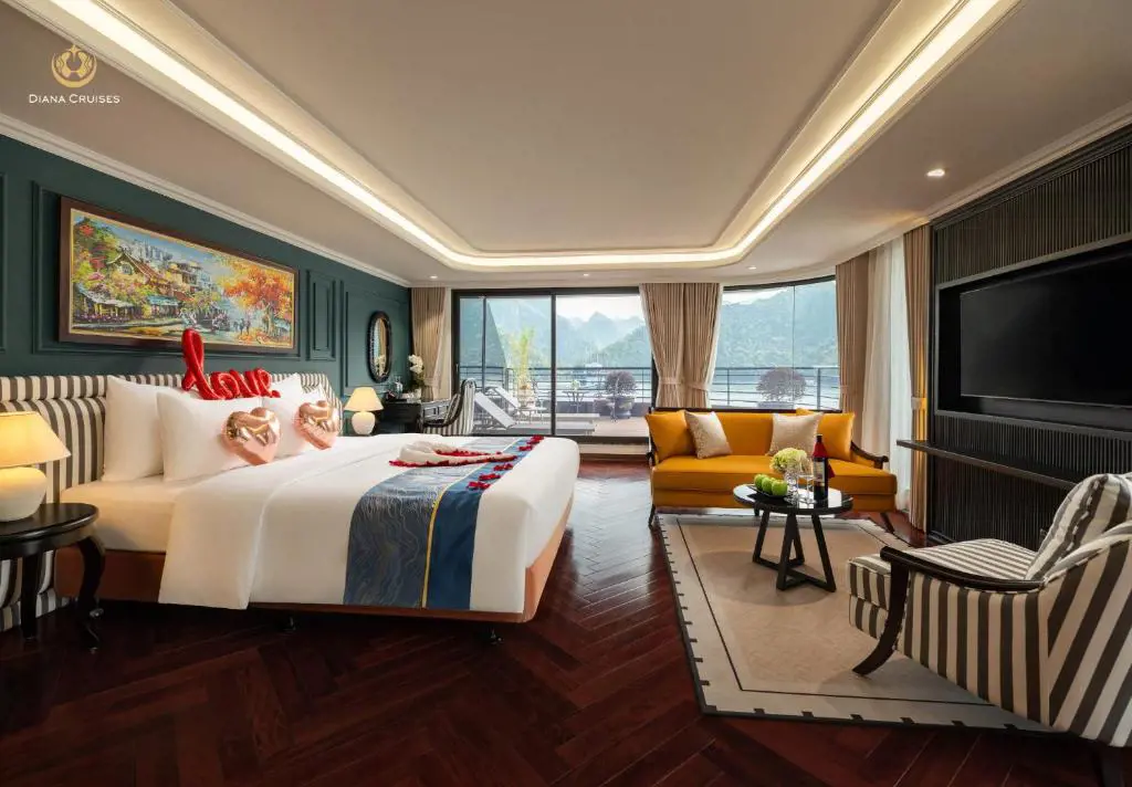 Luxurious cruise ship suite with a romantically decorated bed, mustard yellow sofa, and panoramic floor-to-ceiling windows overlooking scenic bay mountains.