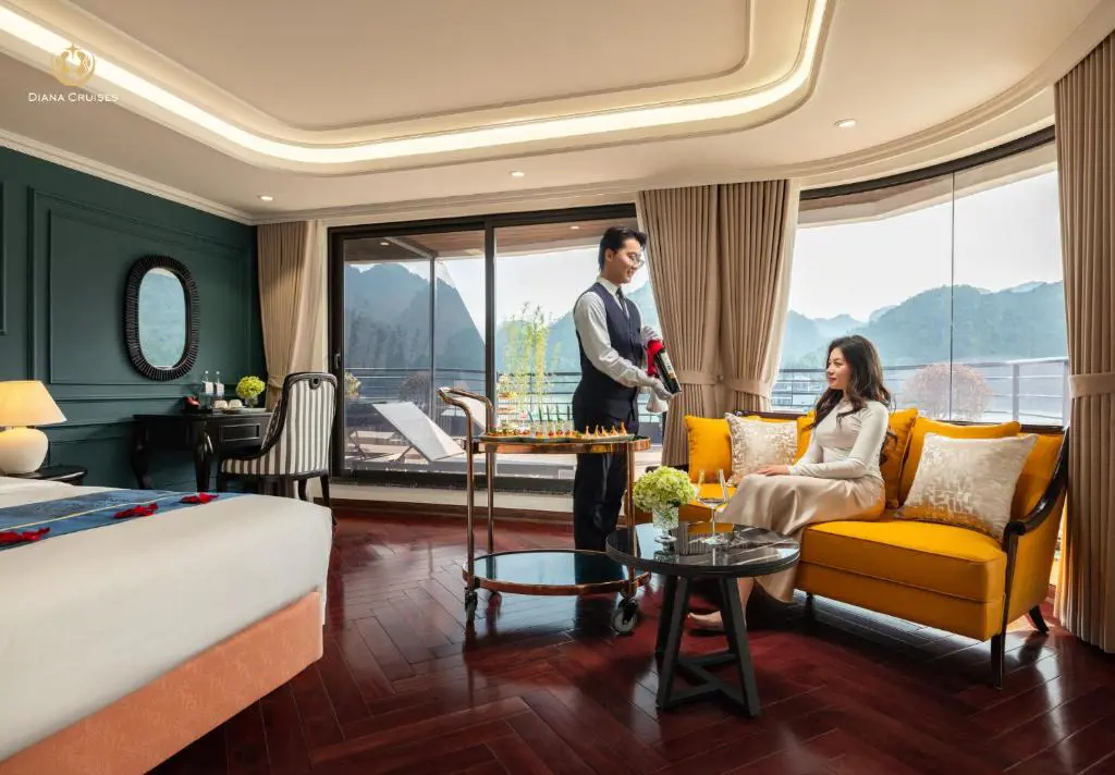 A waiter serving wine to a guest inside a luxurious cruise cabin with panoramic views of limestone karsts.