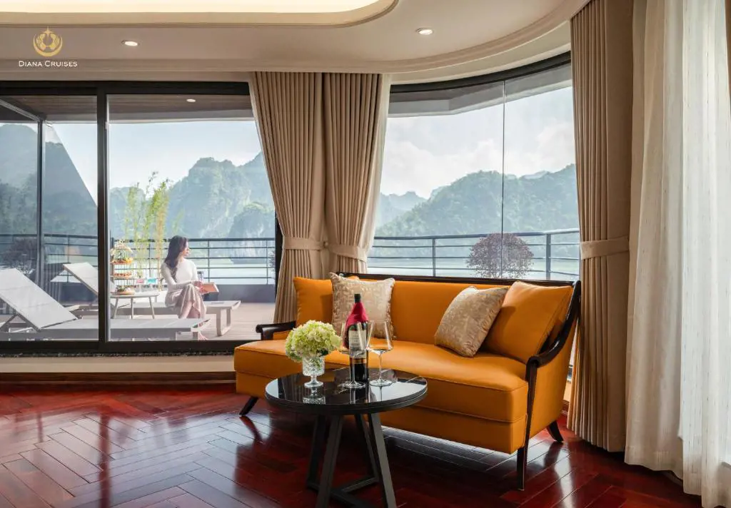 Interior of a luxury cruise suite with an orange sofa, wine set on a small coffee table, floor-to-ceiling windows, and a woman sitting on the private balcony looking at limestone mountains.