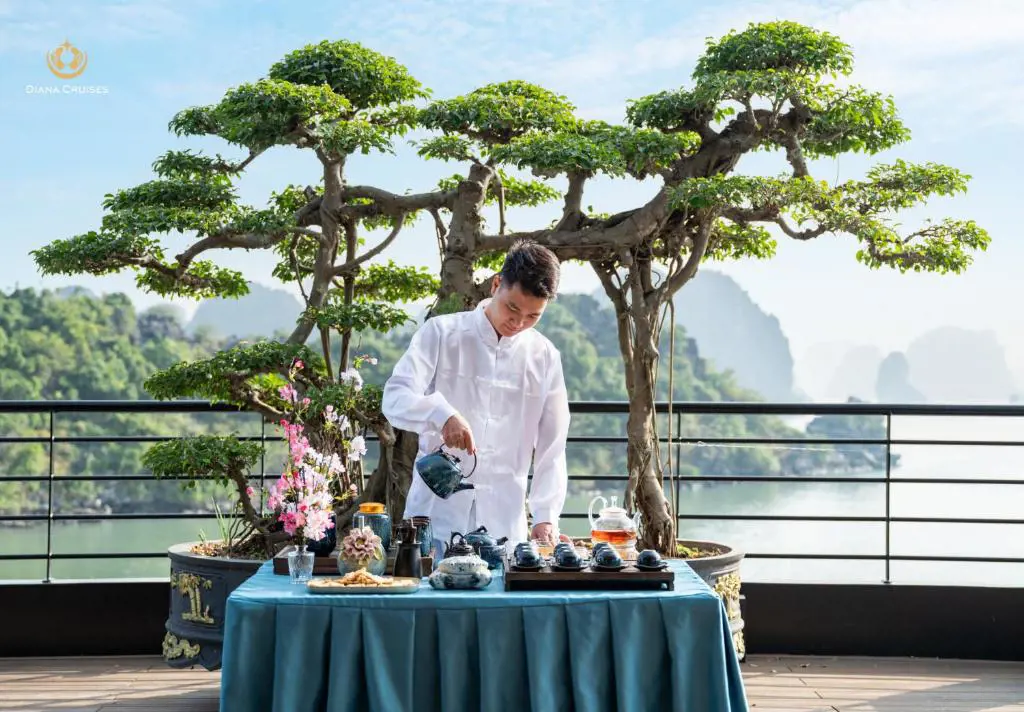 A man in a white shirt pouring tea at a decorated table on a cruise deck in front of a bonsai tree and limestone karst scenery.