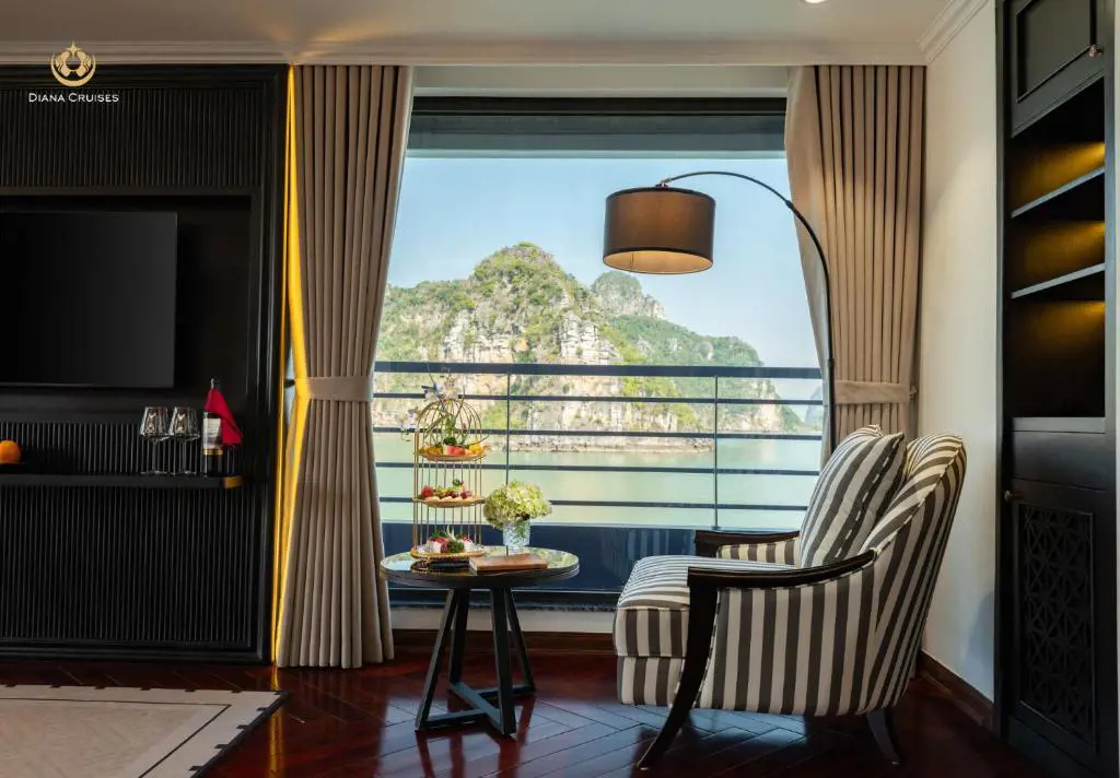 Luxury cruise cabin interior with a striped armchair, afternoon tea set on a table, and a large window showing limestone karst mountains.