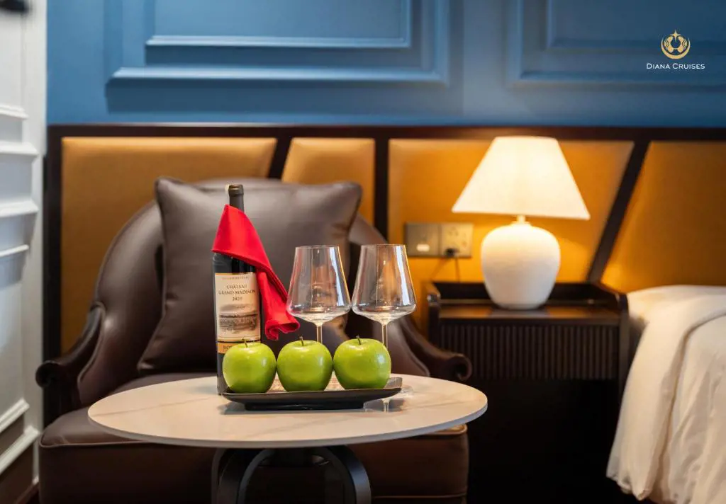 A bottle of wine, two wine glasses, and three green apples on a table inside a luxury cruise cabin suite.