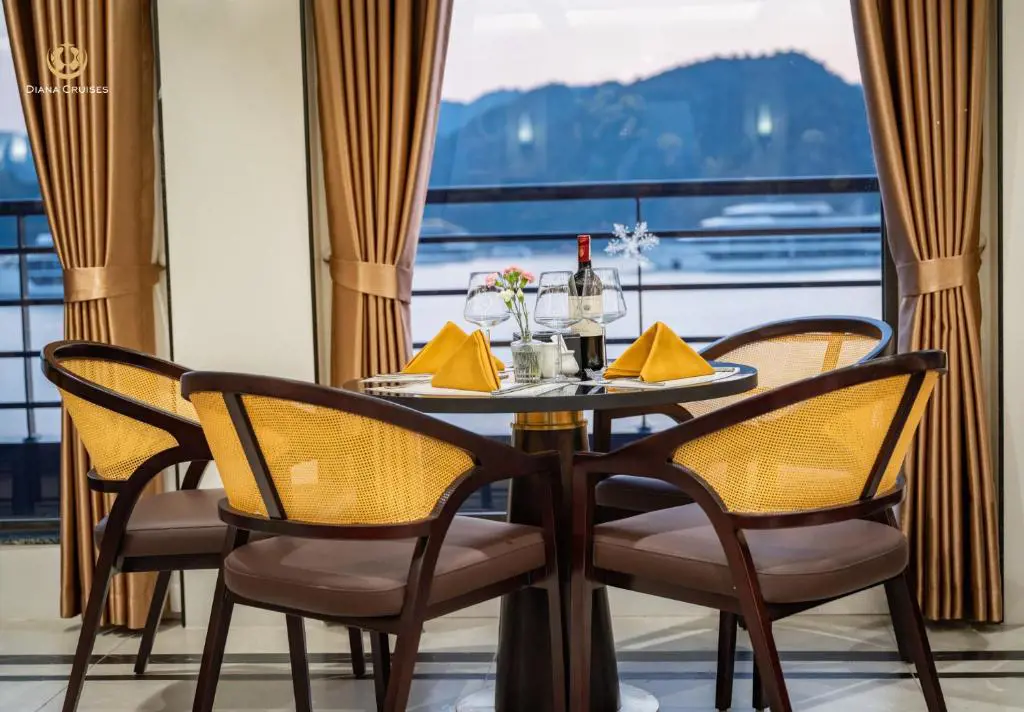 Bàn ăn bốn ghế xếp xung quanh chai rượu vang và ly thủy tinh bên cửa sổ du thuyền Diana Cruises nhìn ra vịnh biển