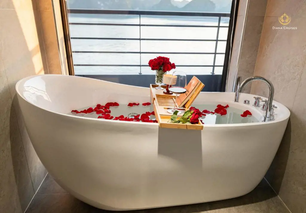 A white freestanding bathtub with red rose petals, a glass of wine on a tray, and a scenic sea view through a window.