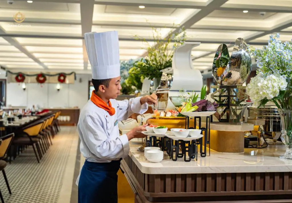 Đầu bếp mặc đồng phục trắng và đội mũ cao đang bài trí món ăn trên quầy buffet sang trọng của du thuyền Diana Cruises.