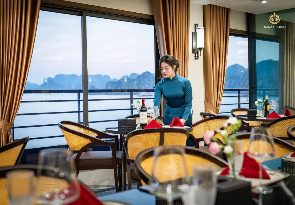 Nữ nhân viên mặc áo dài xanh đang chuẩn bị bàn ăn trong nhà hàng du thuyền hướng nhìn ra cảnh biển núi đá vôi