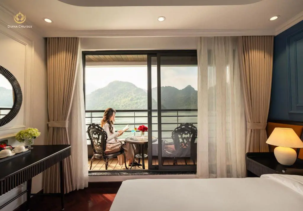 A woman sitting on an ornate chair reading a book on a cruise cabin private balcony with mountains in the background.