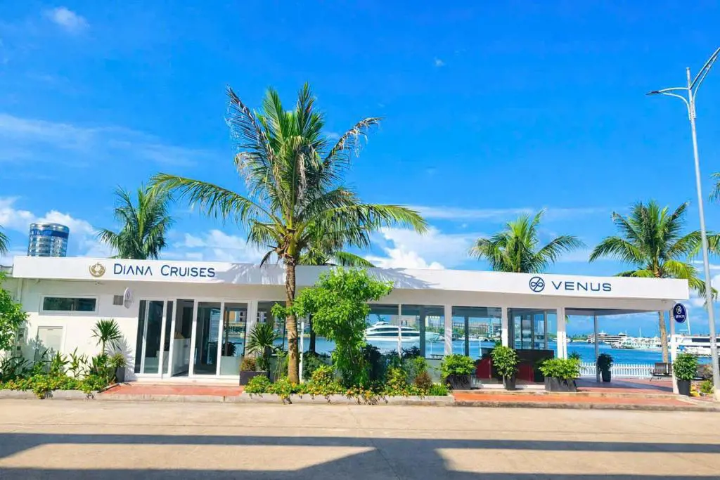 Single-story white terminal building with Diana Cruises and Venus logos, palm trees in front, and boats docked in the background water under a clear blue sky.