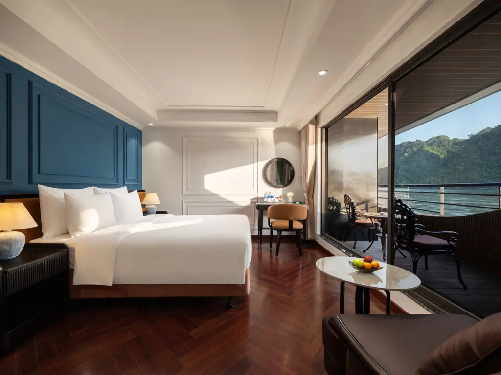 Interior of the Diana Junior Suite cabin with a white bed, blue wall accent, dark wooden floor, and sliding glass balcony doors showing a water and mountain view.