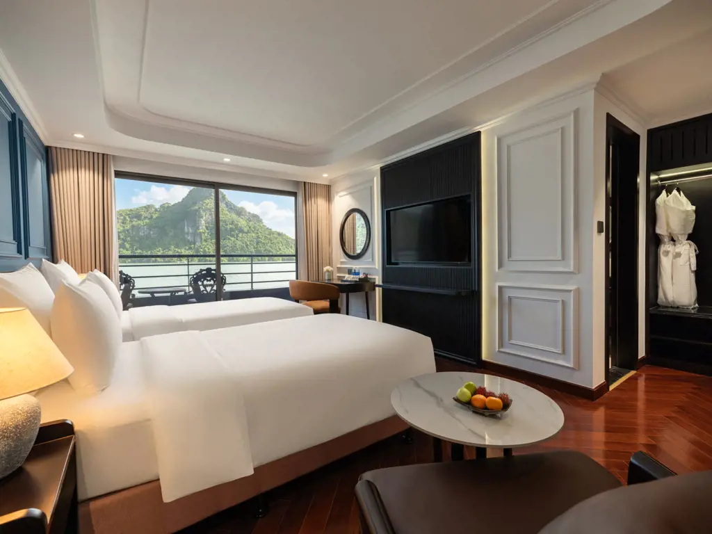 Luxury cruise cabin interior with twin beds, dark wood accents, TV, and large glass balcony door displaying scenic mountain water views.