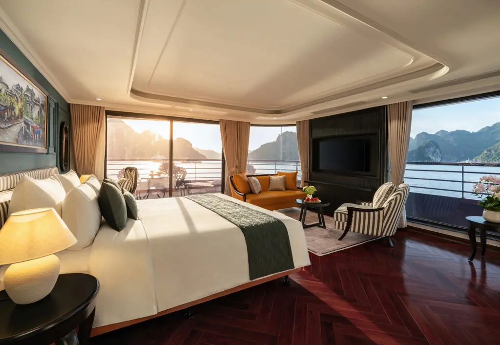 Spacious luxury suite bedroom featuring a large bed, orange sofa, television, and floor-to-ceiling windows showing limestone karst mountains in Ha Long Bay.