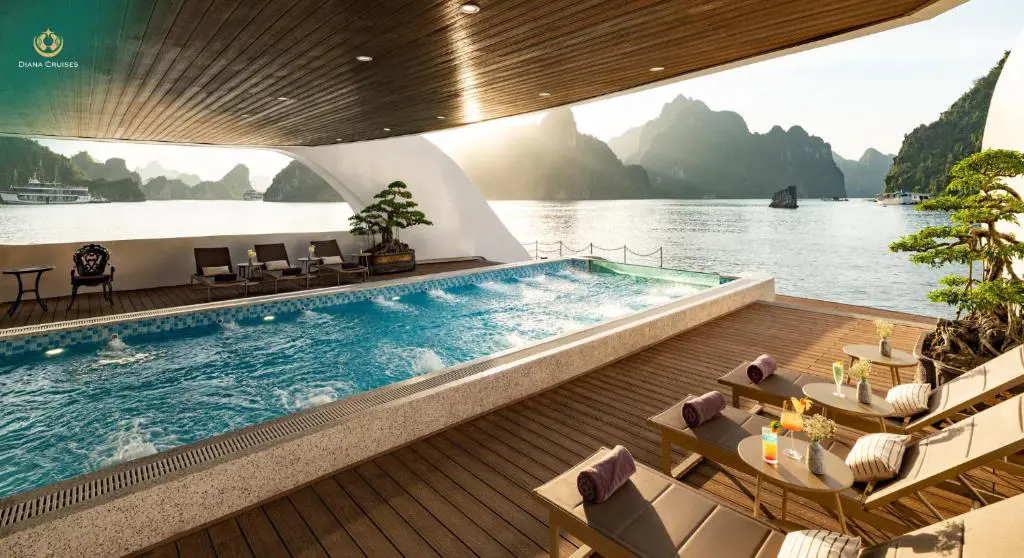 An outdoor cruise ship pool deck featuring lounge chairs, bonsai plants, and a swimming pool framing a panoramic view of limestone island karsts in Ha Long Bay.