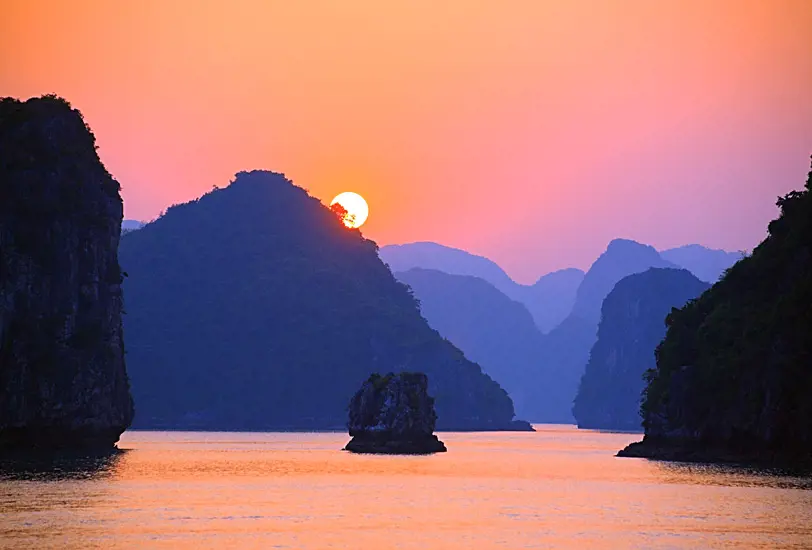 The sun setting behind silhouetted limestone karst peaks over tranquil water illuminated by an orange and purple sky.