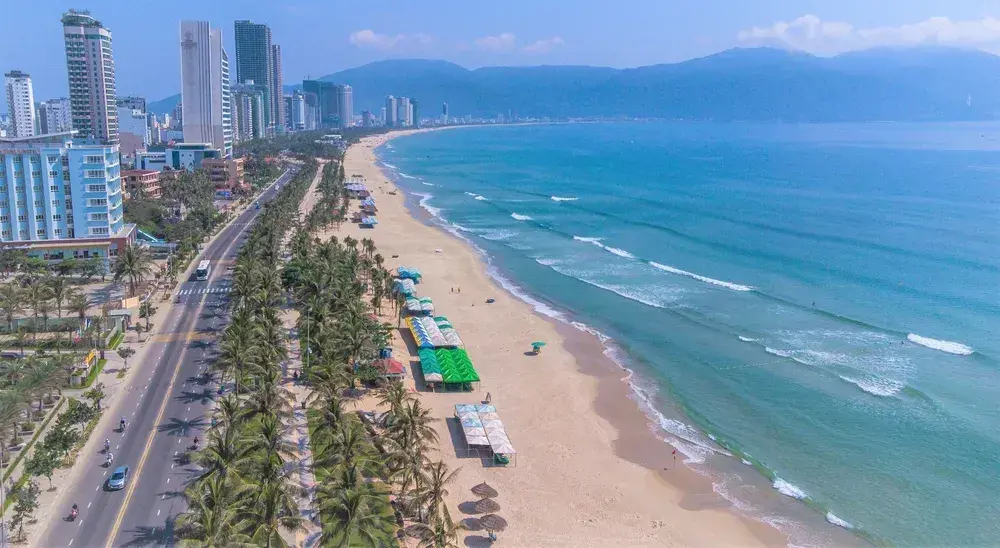 Aerial view of My Khe Beach in Da Nang, featuring a sandy coastline, turquoise ocean, palm-lined street, and tall city buildings.