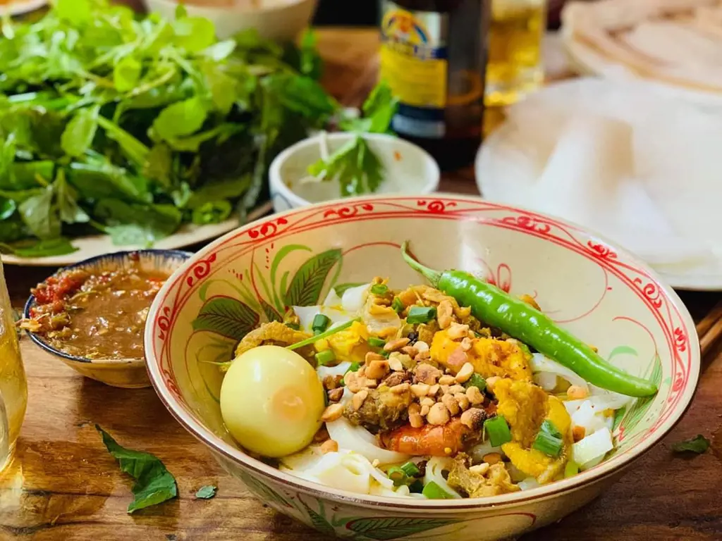 A bowl of Mi Quang noodles topped with a boiled egg, peanuts, scallions, and a whole green chili pepper on a wooden table with fresh herbs.