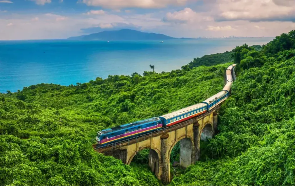 A blue and red passenger train travels across an old stone arch railway bridge amid dense green forest next to a blue ocean.
