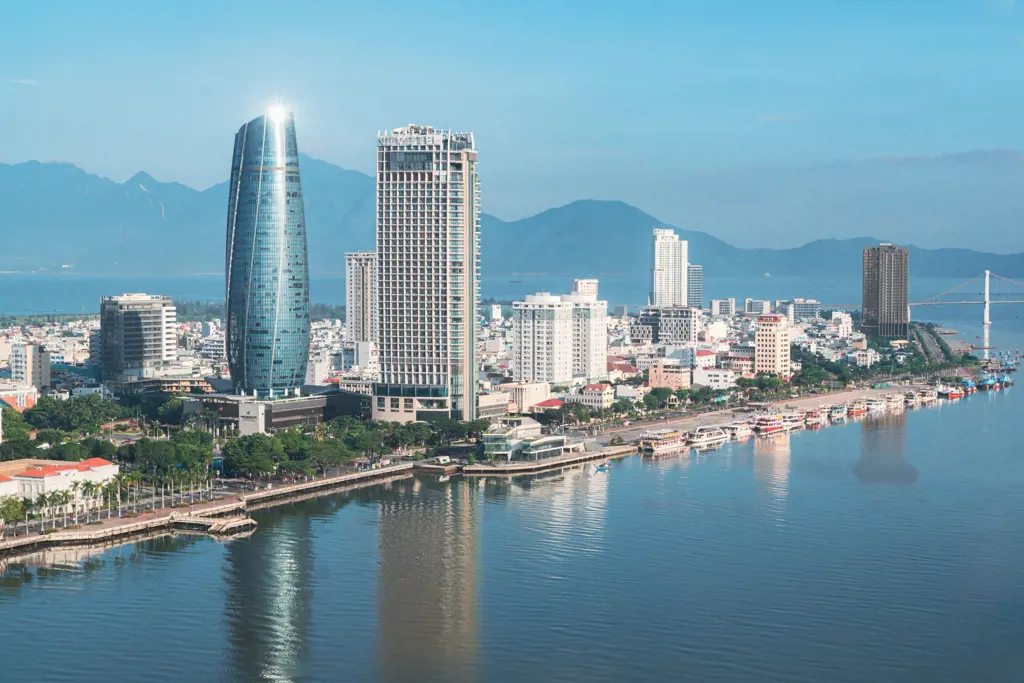 Panoramic view of modern skyscrapers along the Han River in Da Nang, Vietnam, with mountains and a bridge in the background.