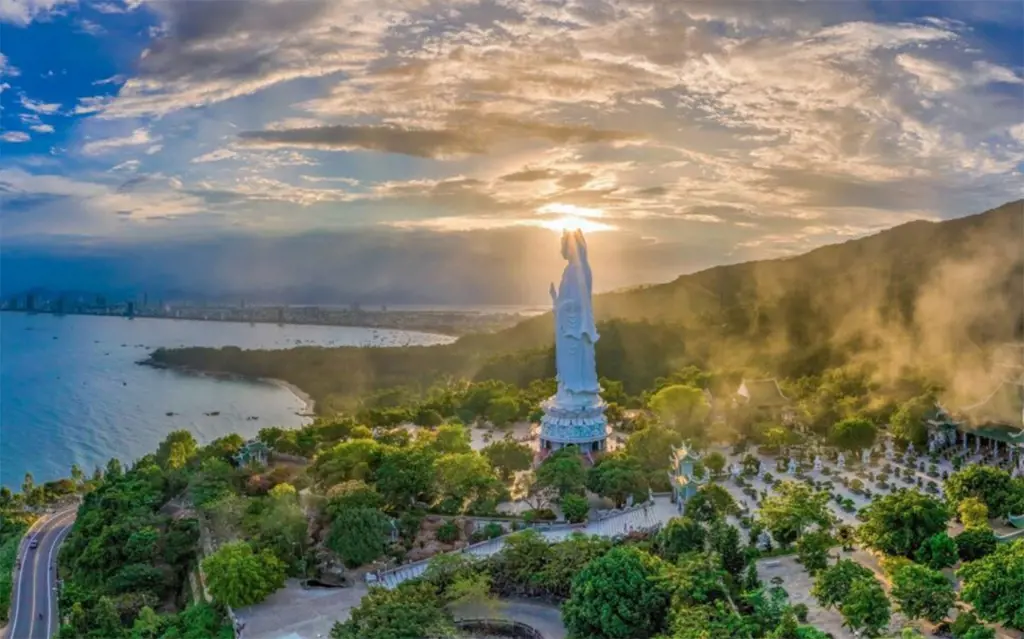Ảnh chụp từ trên cao tượng Quan Âm trắng lớn trong khuôn viên Chùa Linh Ứng trên sườn đồi ven biển, dưới bầu trời nhiều mây và nắng thấp.