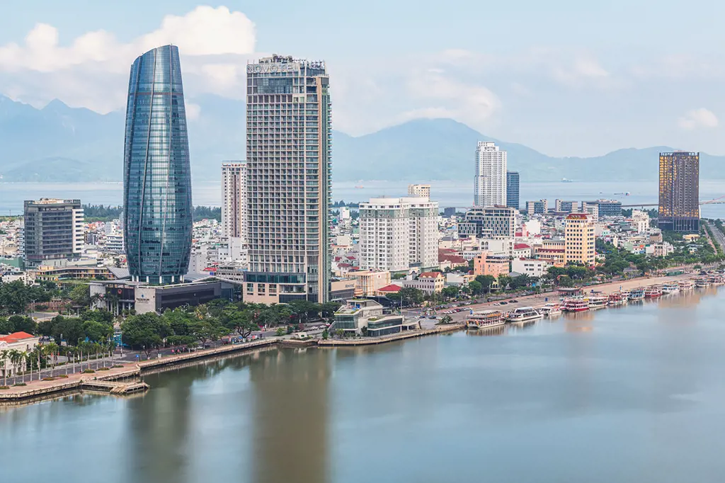Toàn cảnh các tòa nhà cao tầng ven Sông Hàn ở Đà Nẵng, nổi bật với tháp kính bo tròn, bến thuyền và dãy núi phía xa dưới trời sáng. – See Da Nang from the Han River