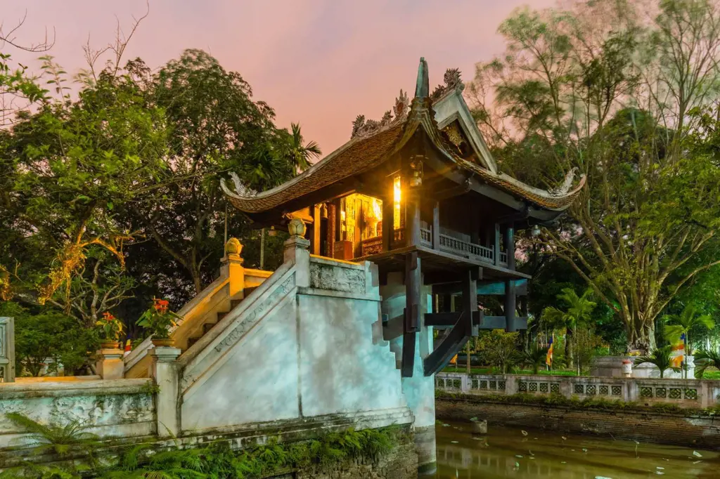 The illuminated One Pillar Pagoda in Hanoi, Vietnam, standing on a stone pillar above a pond surrounded by lush trees under a pink twilight sky.