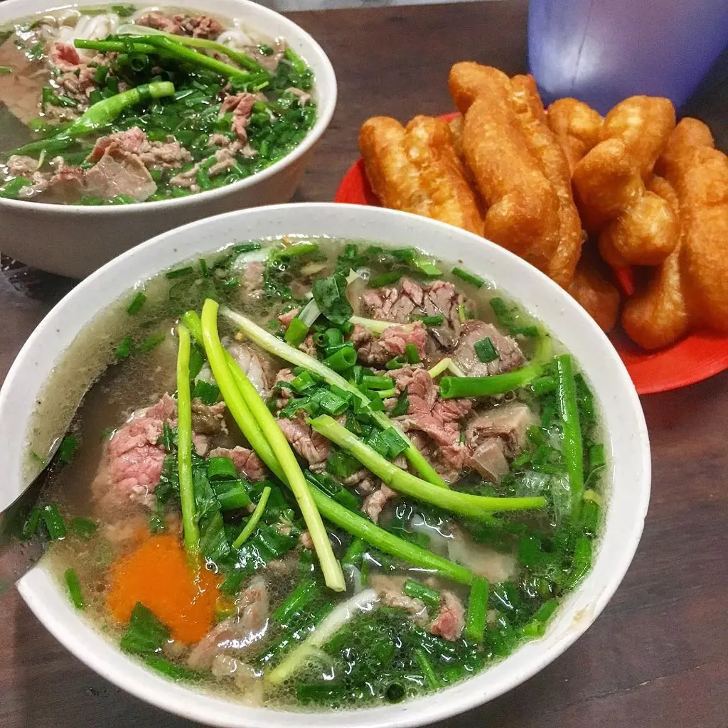 Two bowls of Vietnamese beef noodle soup garnished with abundant green scallions and sliced beef, served alongside a plate of golden fried dough sticks.
