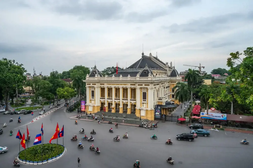 Elevated view of the yellow Hanoi Opera House with motorbikes and cars navigating the surrounding street square in Hanoi, Vietnam.