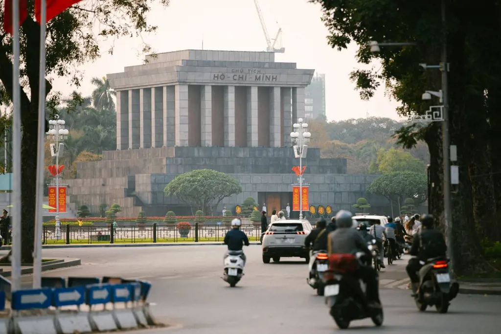 Motorcyclists driving down a street in Hanoi with the large stone Ho Chi Minh Mausoleum visible in the background.