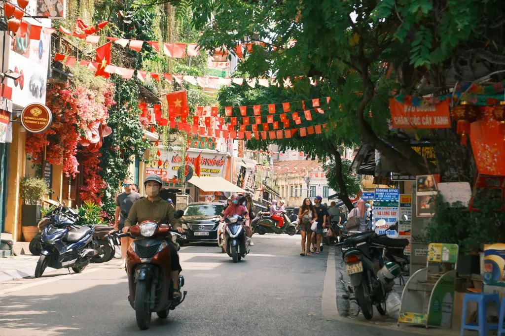 Motorcyclists and pedestrians on a street in Hanoi decorated with red Vietnamese flags.