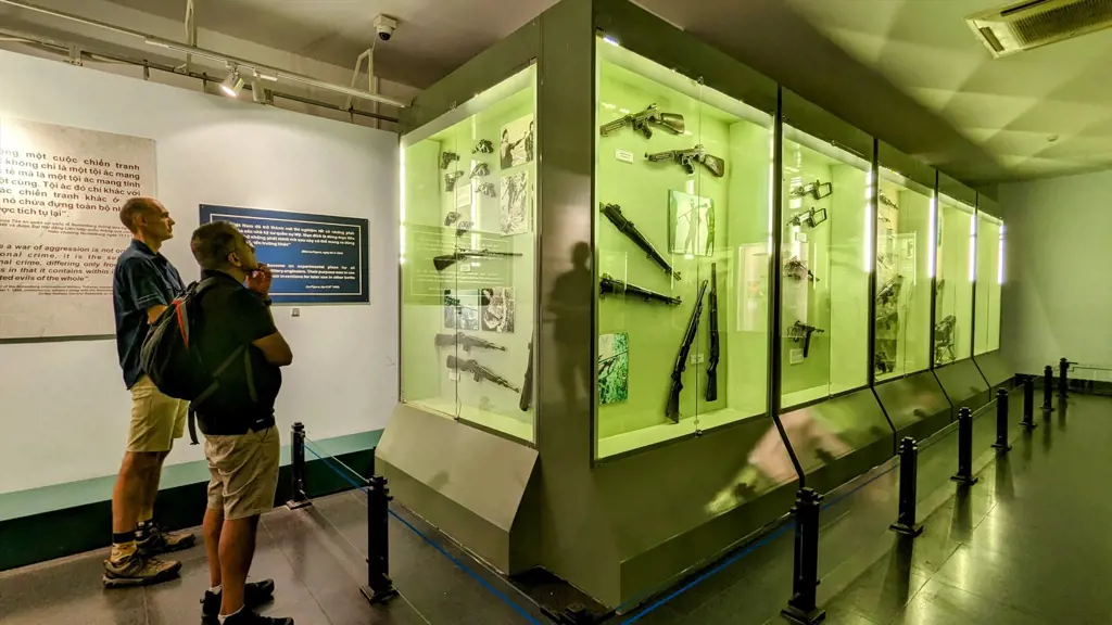 Two tourists standing in front of green-lit display cases containing historical guns and military weapons inside a museum.