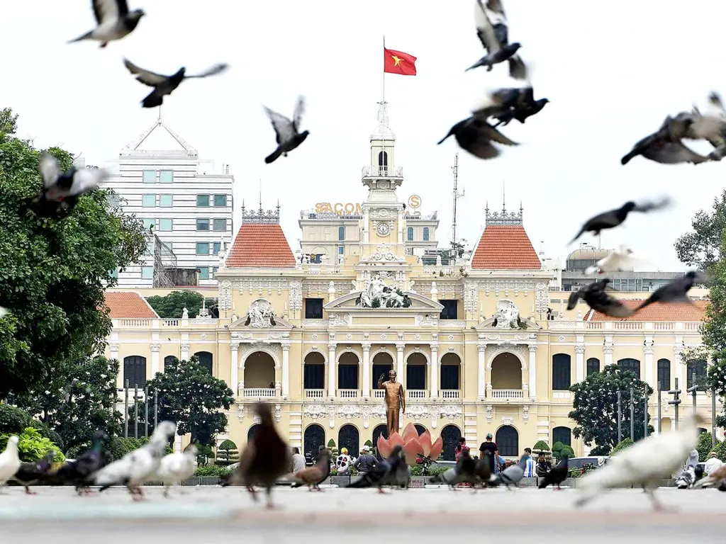 Pigeons flying and gathering on Nguyen Hue Walking Street in front of the historic yellow Ho Chi Minh City Hall building.