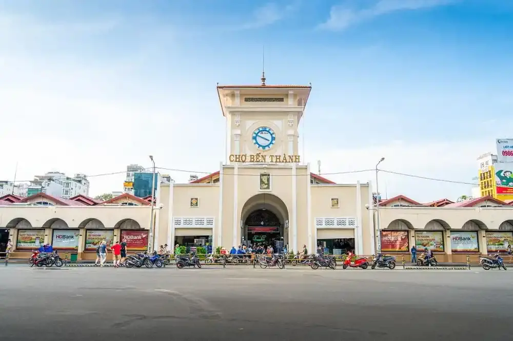 Exterior facade and central clock tower of Ben Thanh Market with pedestrians and motorbikes on the street in Ho Chi Minh City.