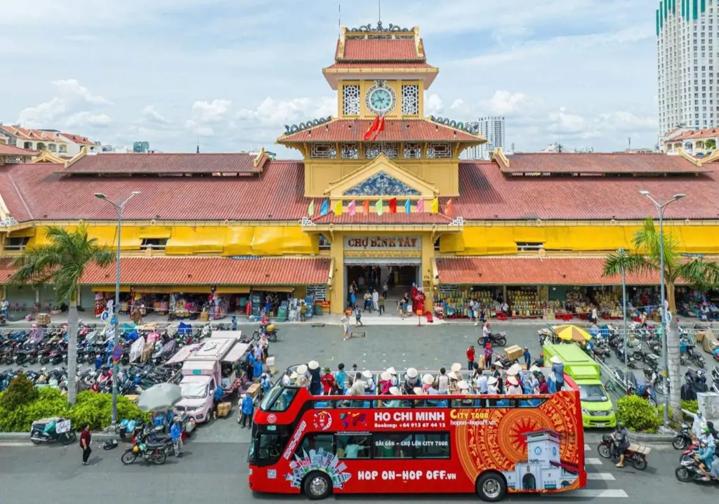 A red open-top double-decker sightseeing bus filled with tourists driving past the yellow front entrance and clock tower of Binh Tay Market.