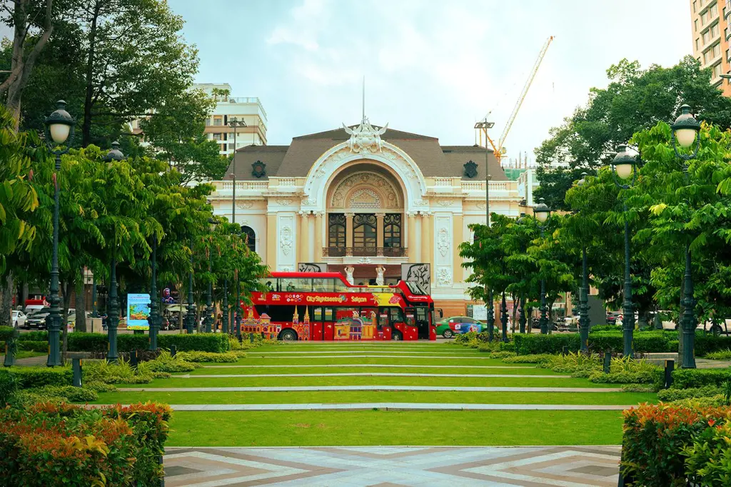 Saigon Opera House seen across a manicured green lawn in Lam Son Square with a red double-decker tour bus parked in front.