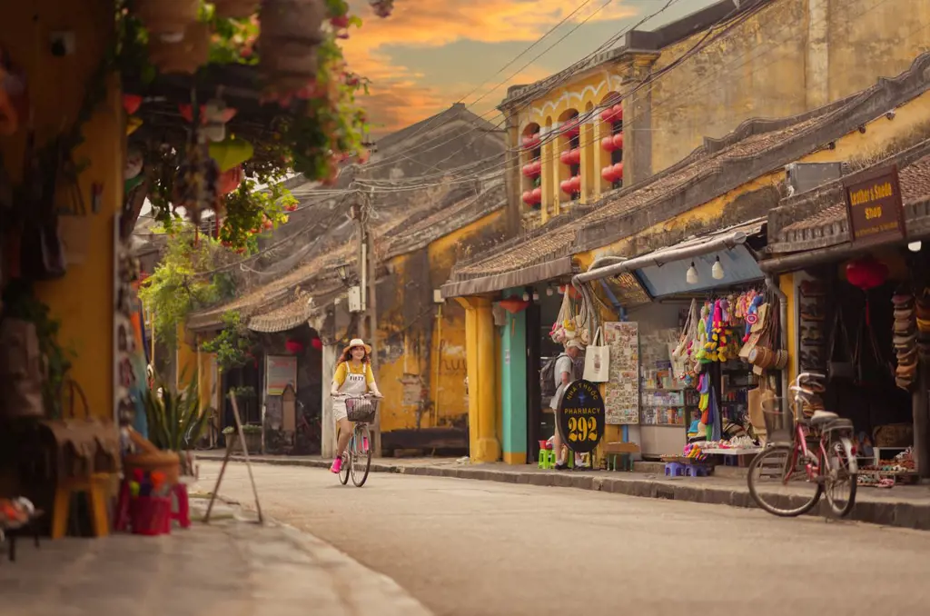 A woman wearing a hat rides a bicycle down a street flanked by historic yellow buildings and local shops in Hoi An Ancient Town under a golden sky.