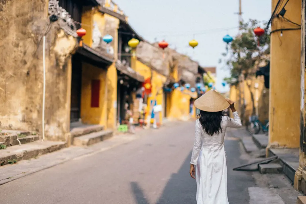 Rear view of a woman in a white Ao Dai and conical hat walking down a street with yellow historic buildings and colorful lanterns in Hoi An.