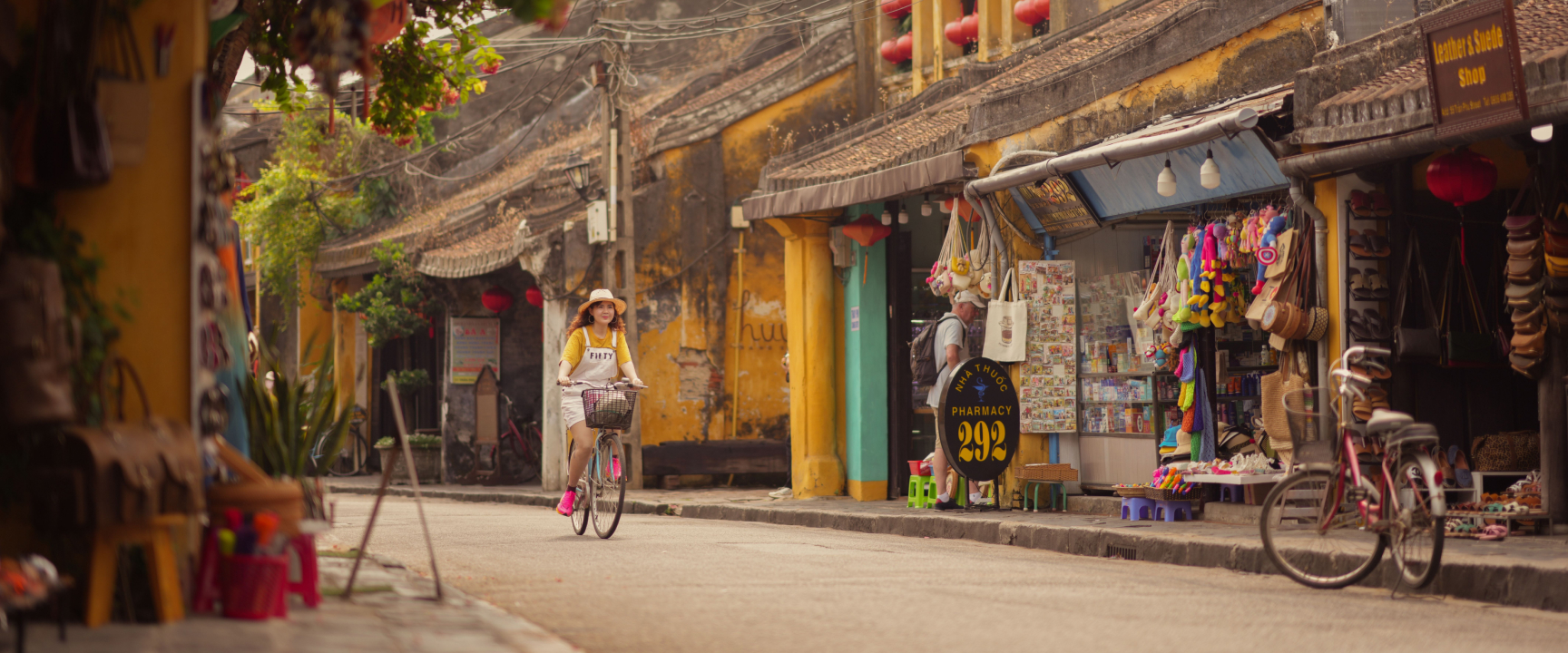 A woman wearing a straw hat rides a bicycle along a historic street lined with yellow buildings and local shops in Hoi An Old Town.