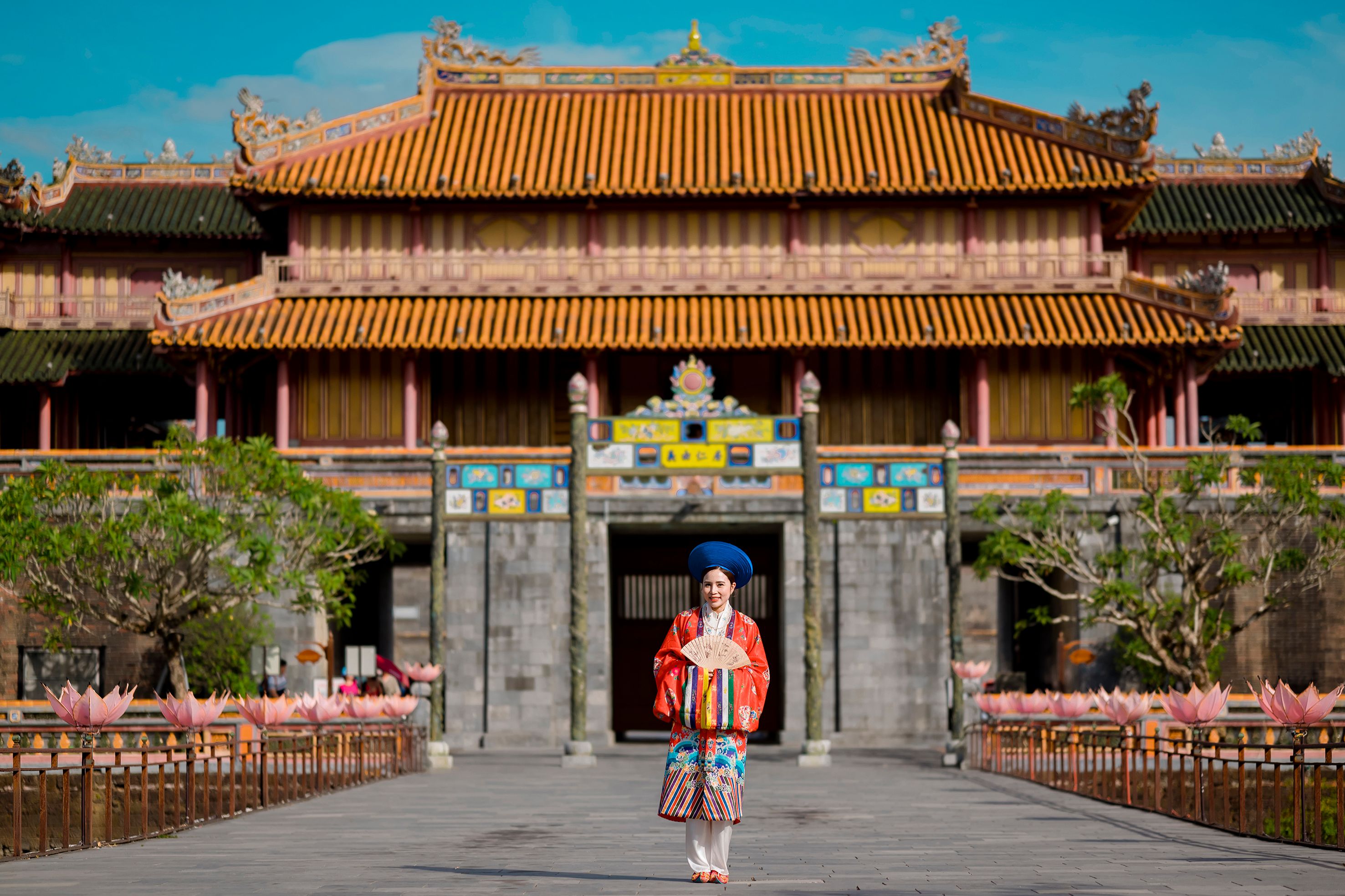 A woman in a red traditional Nhat Binh dress and blue headpiece stands in front of Ngo Mon Gate at the Imperial City in Hue.
