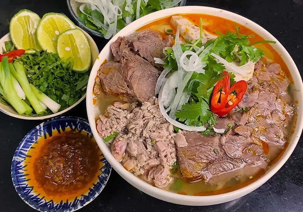 A rich bowl of Bun Bo Hue topped with beef slices, onions, and chili, served with lime wedges and herbs.