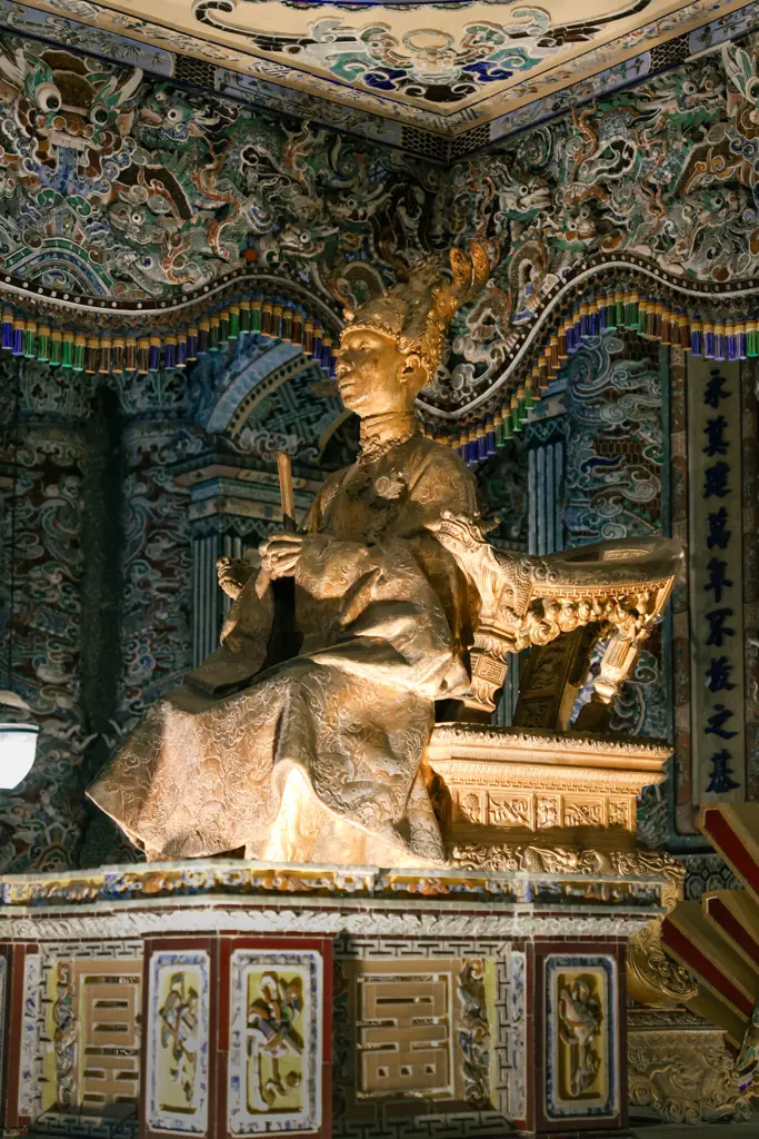 A golden statue of Emperor Khai Dinh seated on a throne inside an intricate mosaic room at Khai Dinh Tomb in Hue, Vietnam.