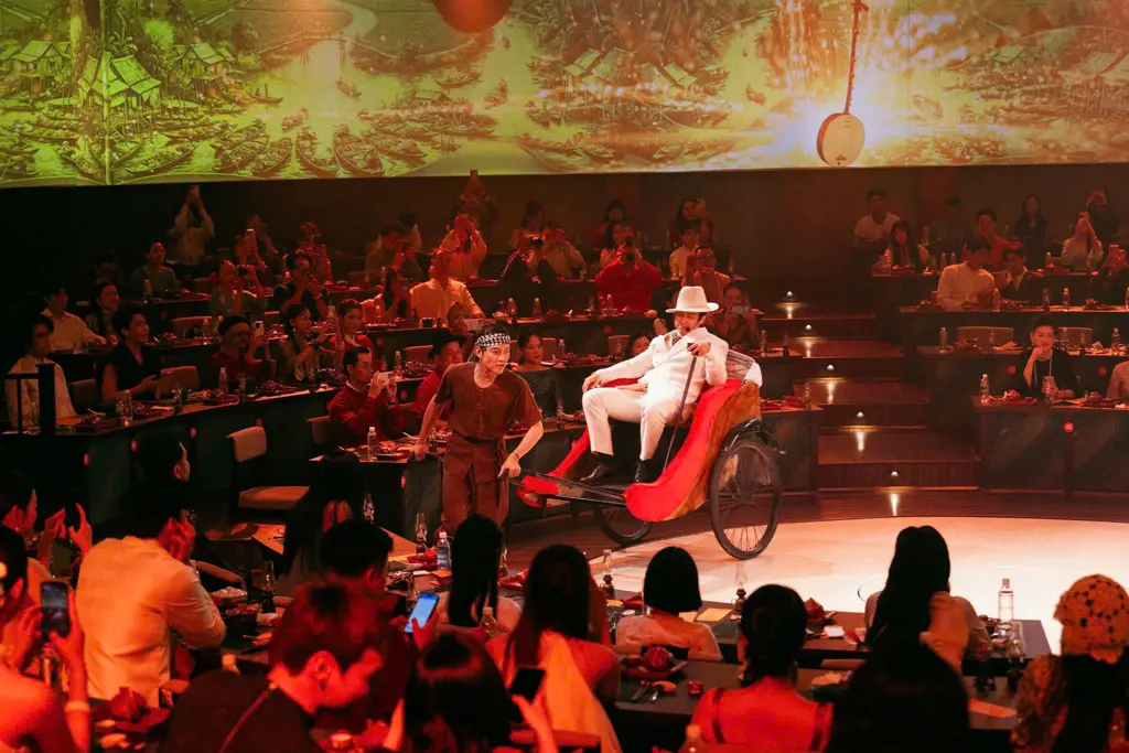 An actor in traditional brown clothing pulls a rickshaw carrying a man in a white suit during a live stage performance.