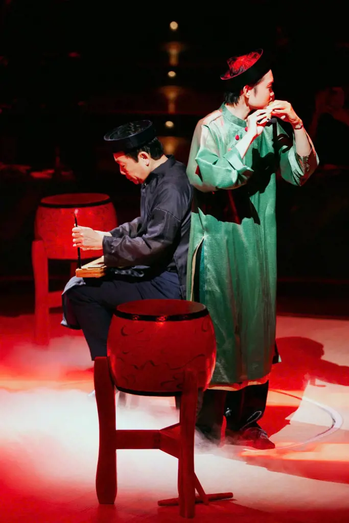 Two musicians in traditional Vietnamese attire play a flute and a stringed instrument on a red-lit stage with low fog and ceremonial drums.
