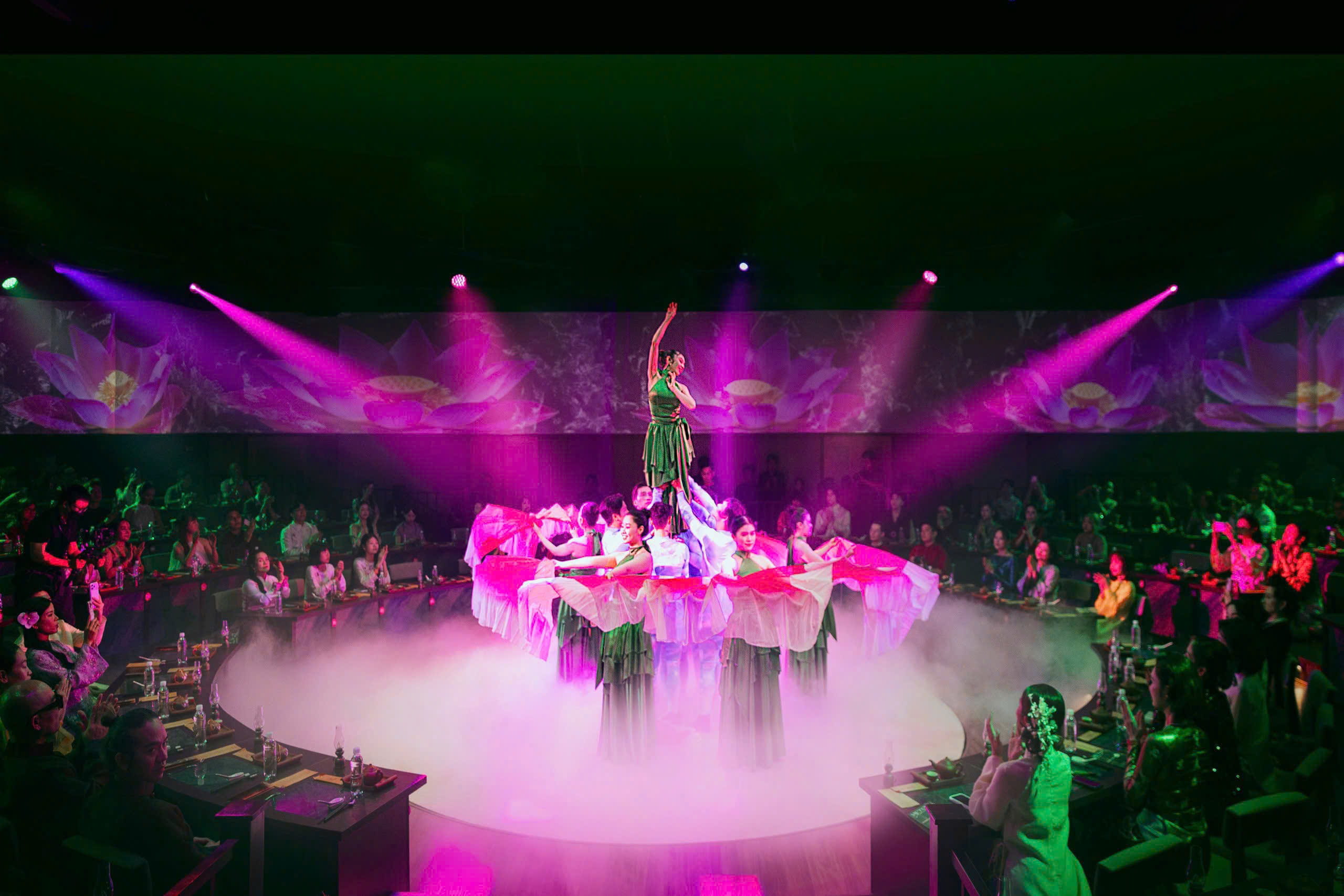 Dancers in green costumes perform on a misty circular stage illuminated by purple and pink spotlights during the Lien Hoa Khoi Thuy show.