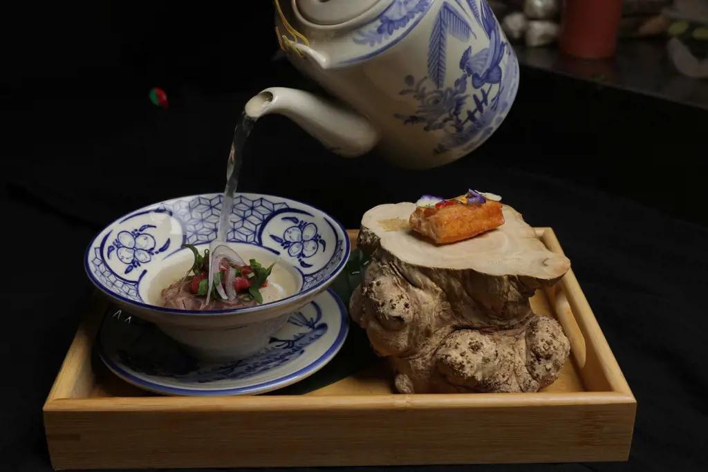 Clear broth pouring from a blue-and-white ceramic teapot into a bowl of sliced meat and herbs on a wooden tray.