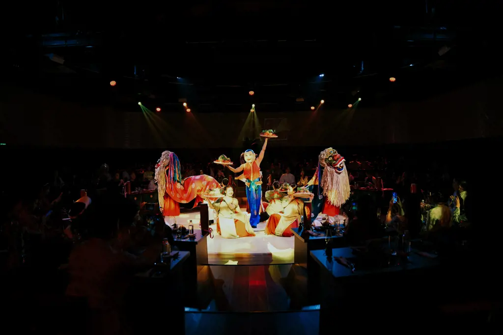 Performers in colorful traditional costumes, a masked character, and lion dance figures on a brightly lit stage in front of an audience.
